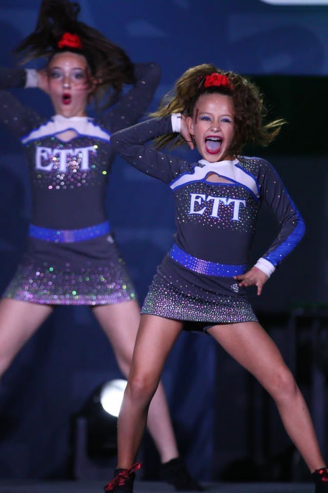 Two cheerleaders in sparkly black uniforms performing on stage, one posing with hands on hips and one mid-move.