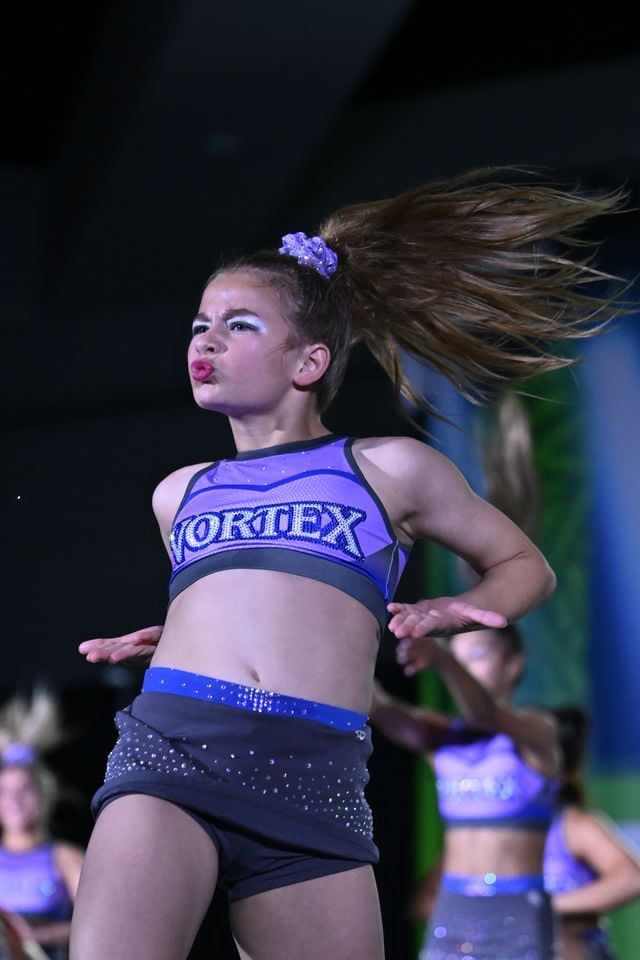 Cheerleader in purple “VORTEX” outfit mid-performance, hair flying, on a dim stage