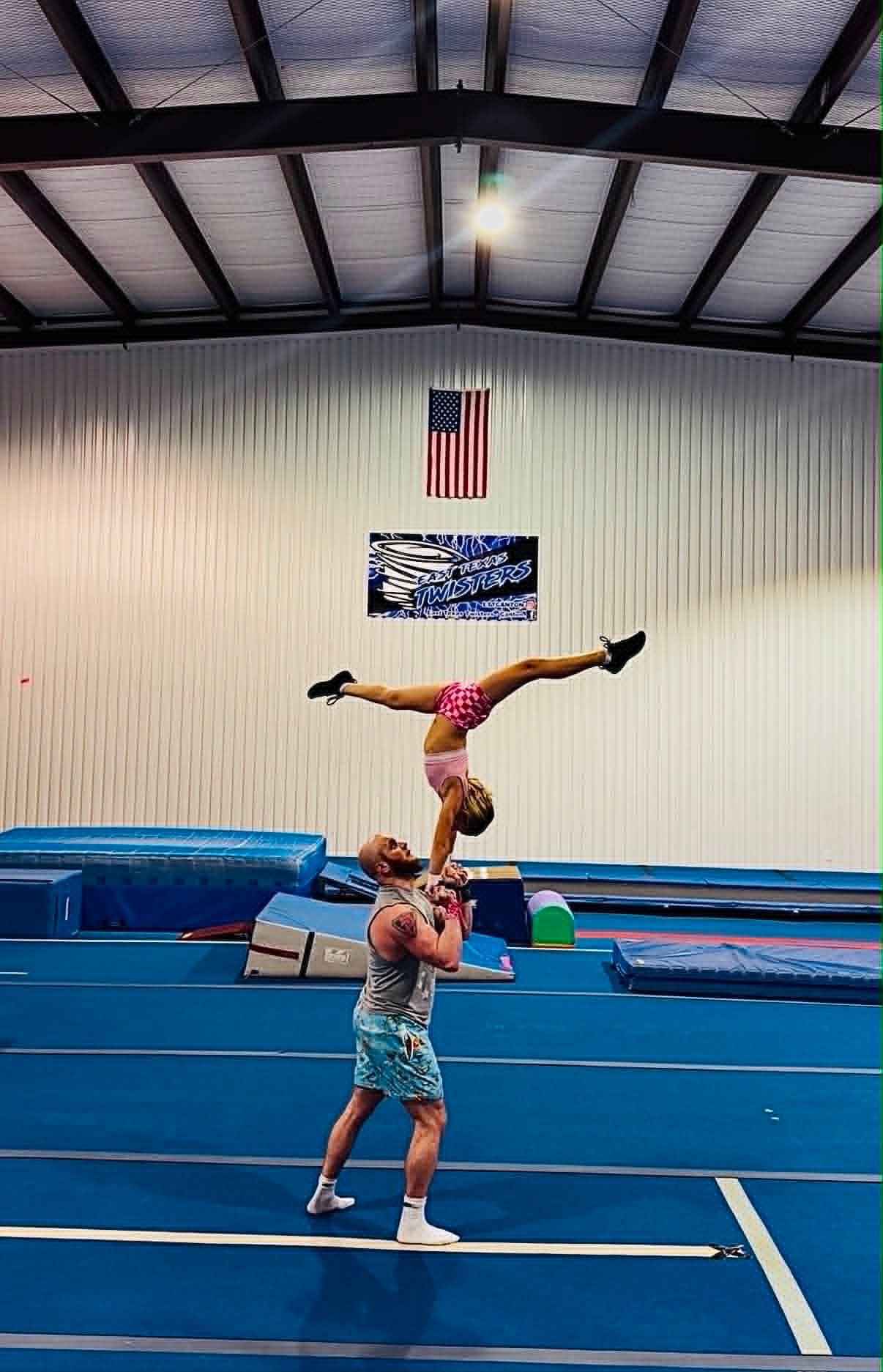 Gymnast balancing on one hand atop another gymnast in a blue training gym