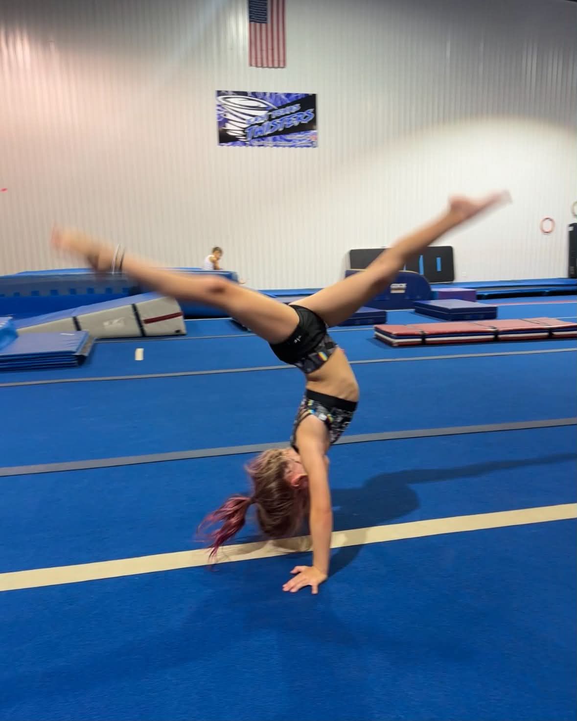 Gymnast performing a one-handed handstand on a blue mat in an indoor gym.