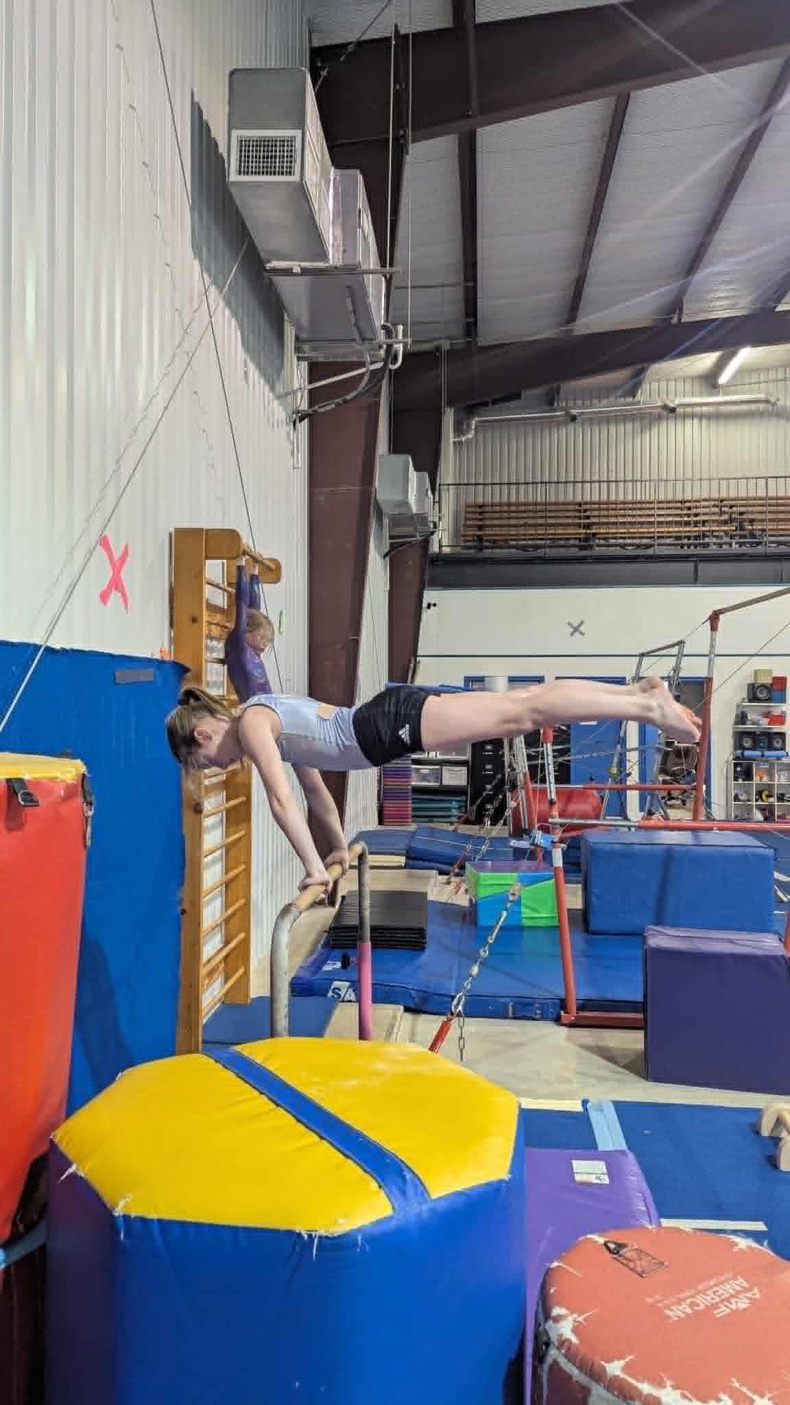 Person balancing horizontally on a gym bar between blue mats in an indoor training facility