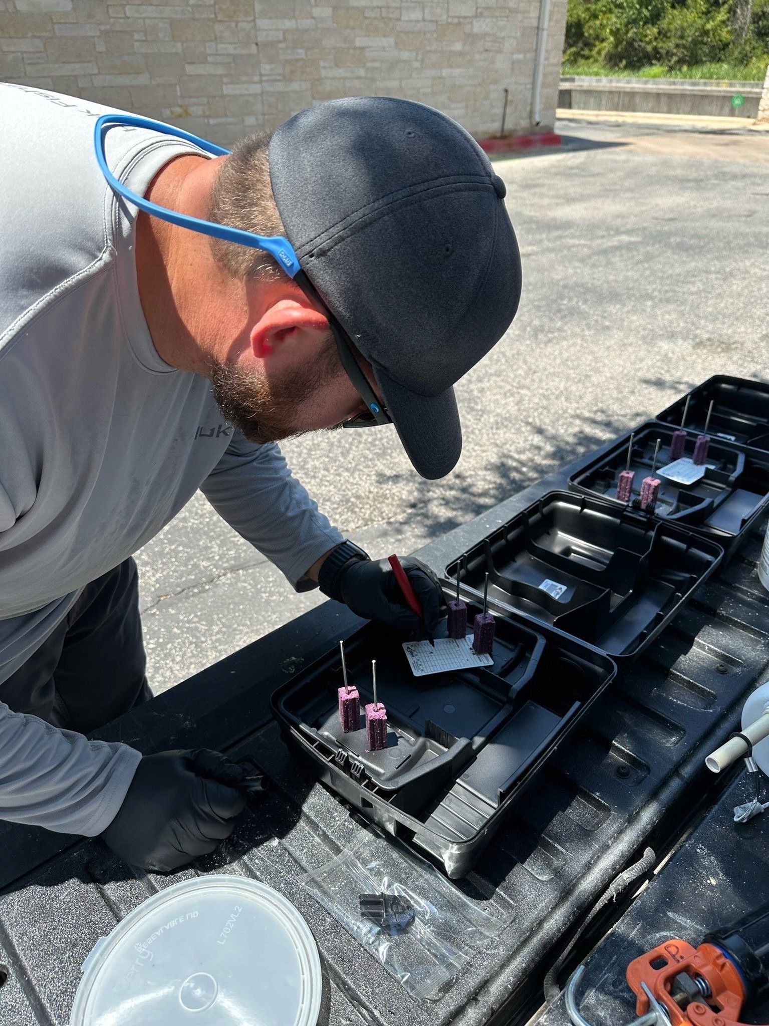 A man wearing a cap and gloves works on a black container, outdoors.