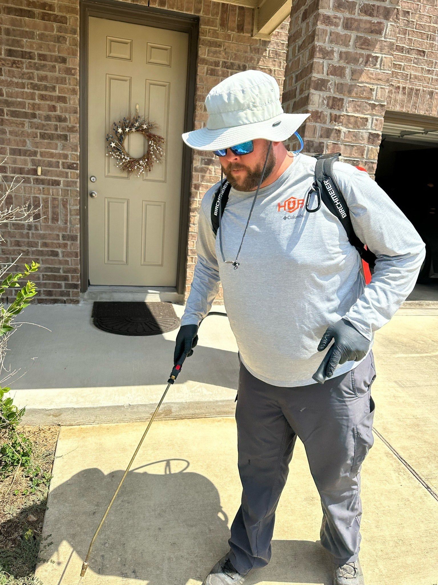Man spraying pesticide on a sidewalk in front of a house. He's wearing a hat, glasses, gloves, and a backpack sprayer.
