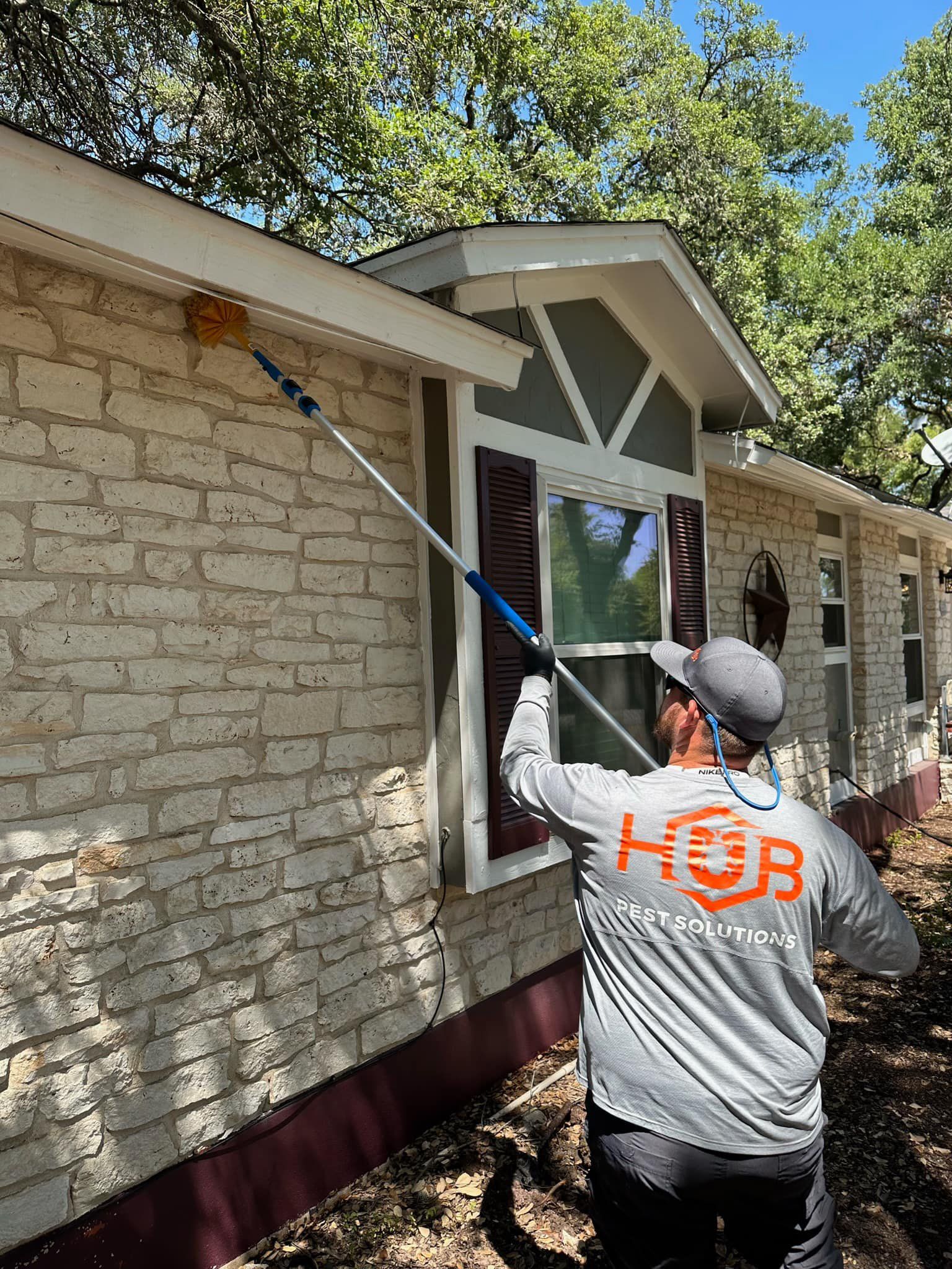Man in gray shirt, cleaning house's edge with a pole brush. Brick wall, window, sunny day.