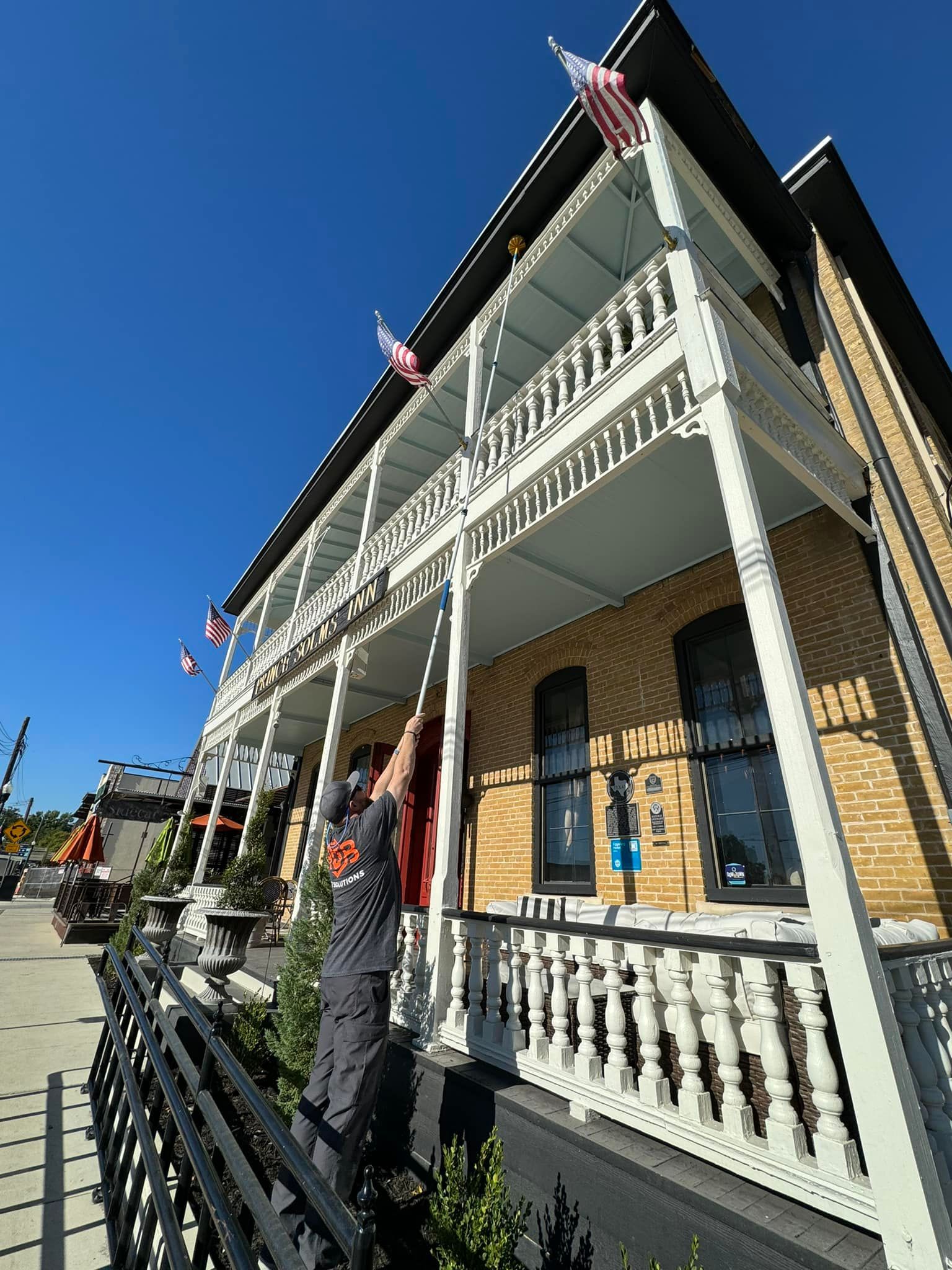 Man adjusting a flag on a two-story building with white trim. American flags on the porch under a blue sky.