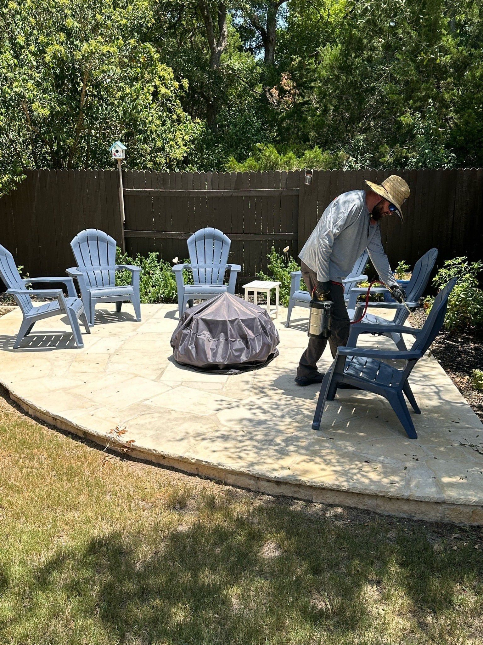 Man arranging chairs around a fire pit on a patio, surrounded by blue chairs, trees and grass.