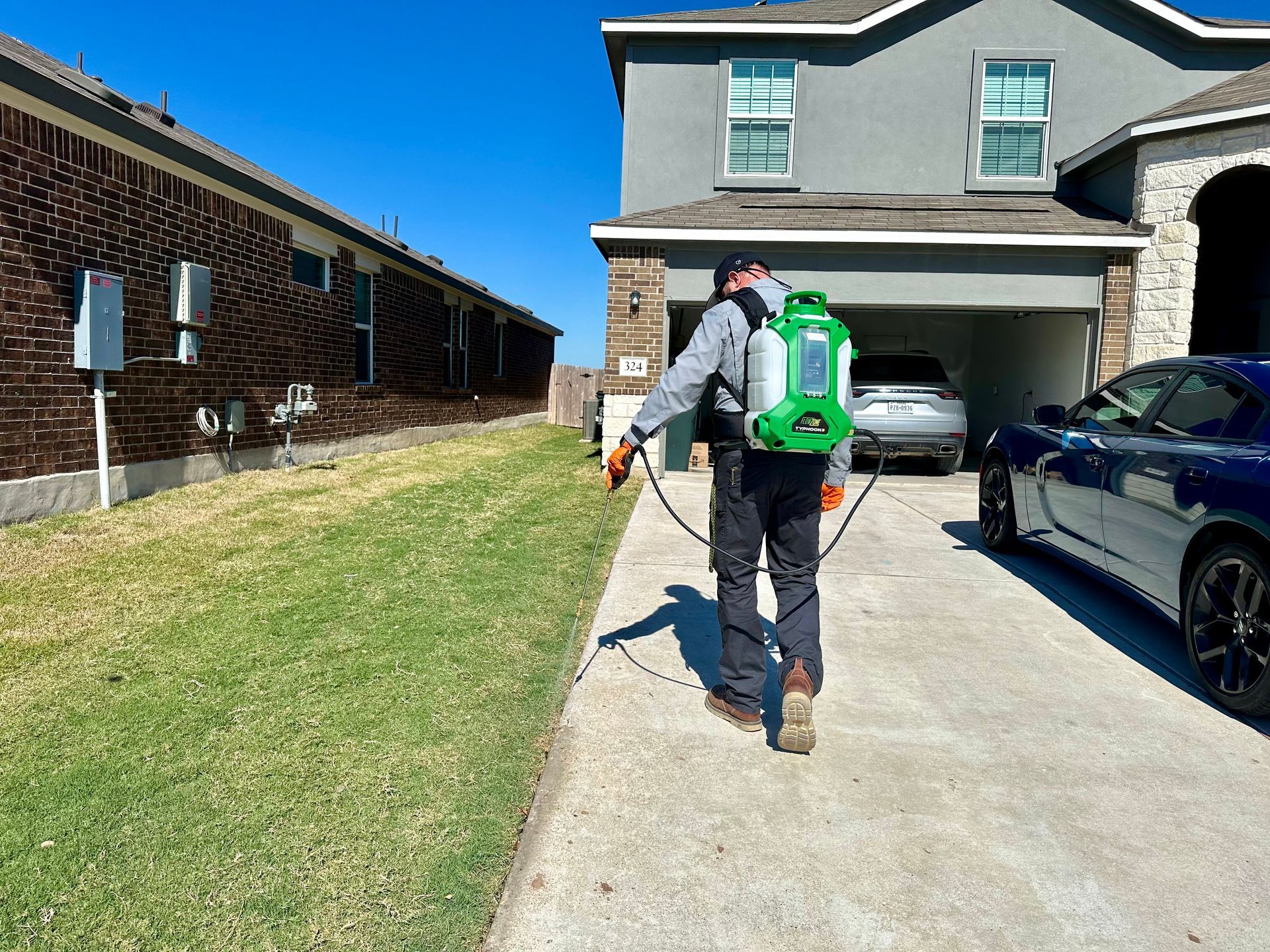 Man spraying lawn with a backpack sprayer next to a house and driveway on a sunny day.