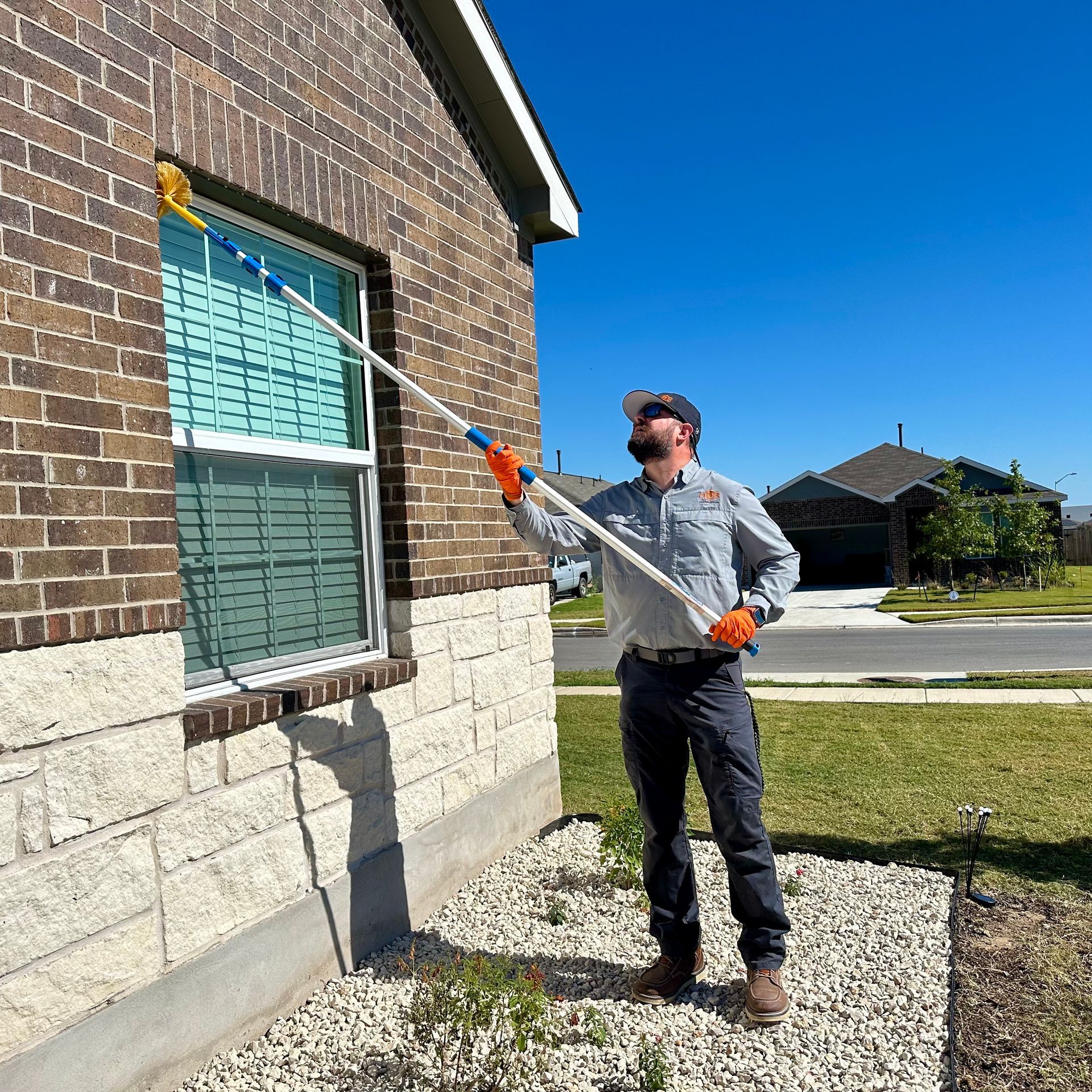 Man cleaning a window on a brick house with a long-handled brush, blue sky overhead.