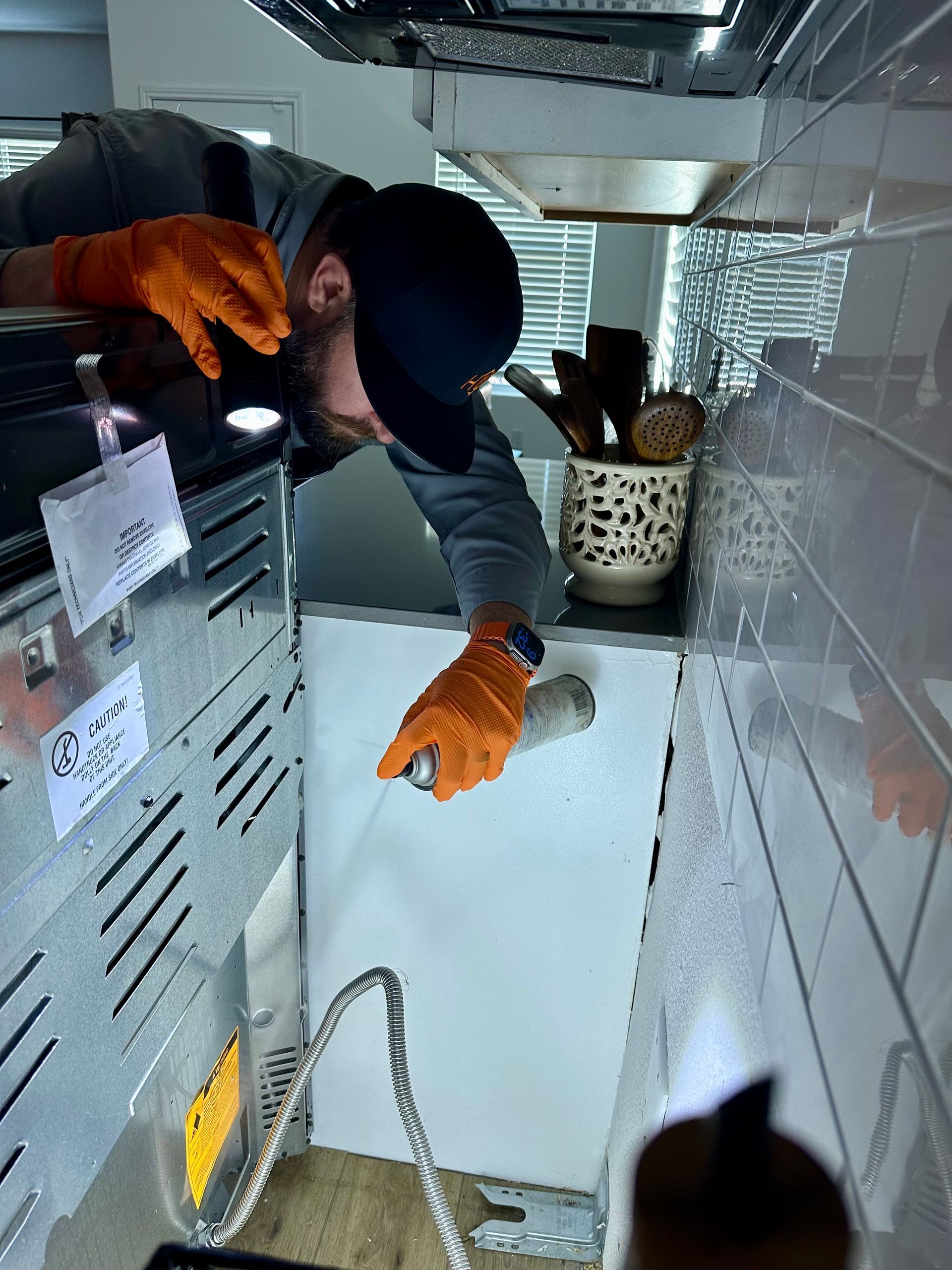 Person sprays beneath a stove in a kitchen, wearing orange gloves and a cap.