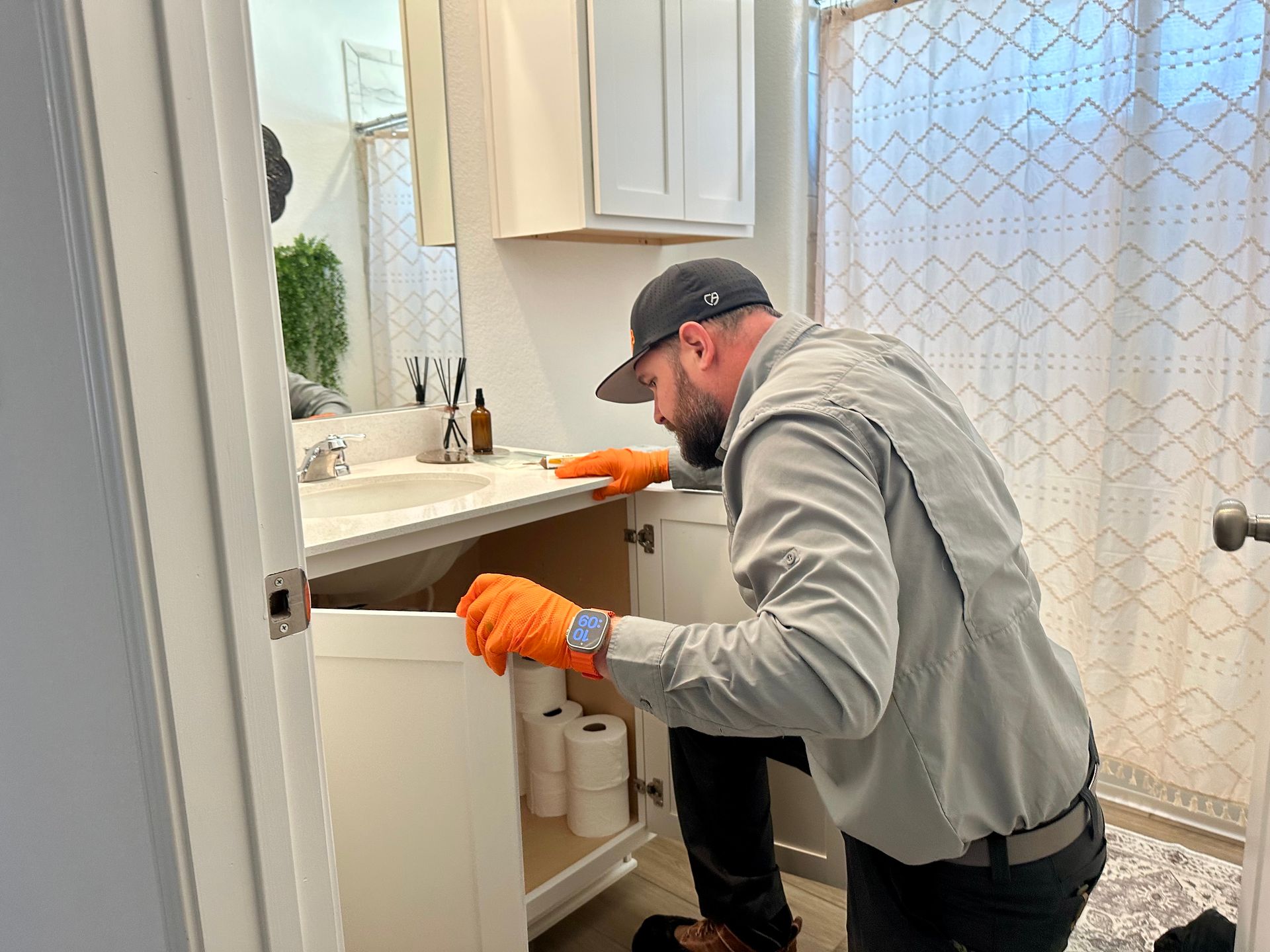 Plumber kneels to work under a bathroom vanity. He wears a hat, gloves, and light shirt in a well-lit space.
