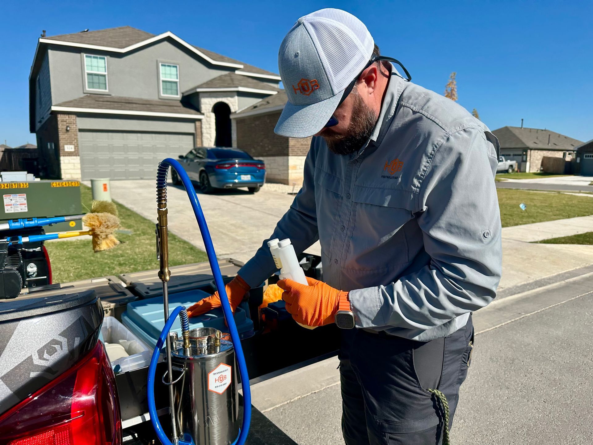 Man in orange gloves, a work shirt, and a cap working on equipment in a truck bed outside a house.