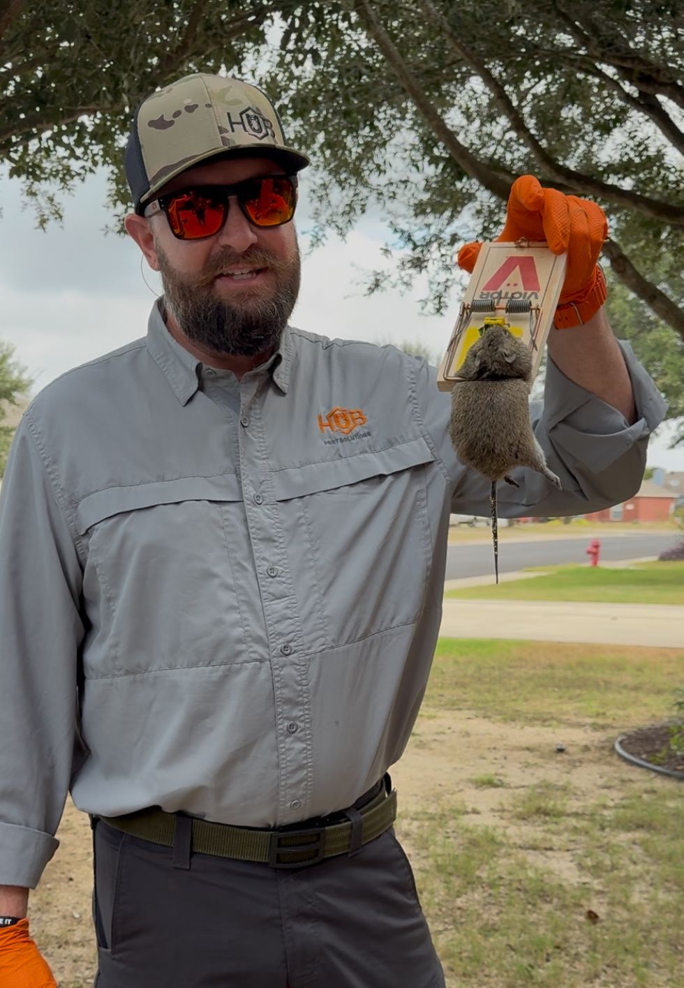 Man holding a mouse trap with a dead mouse. Outdoors, wearing orange gloves and a ball cap.