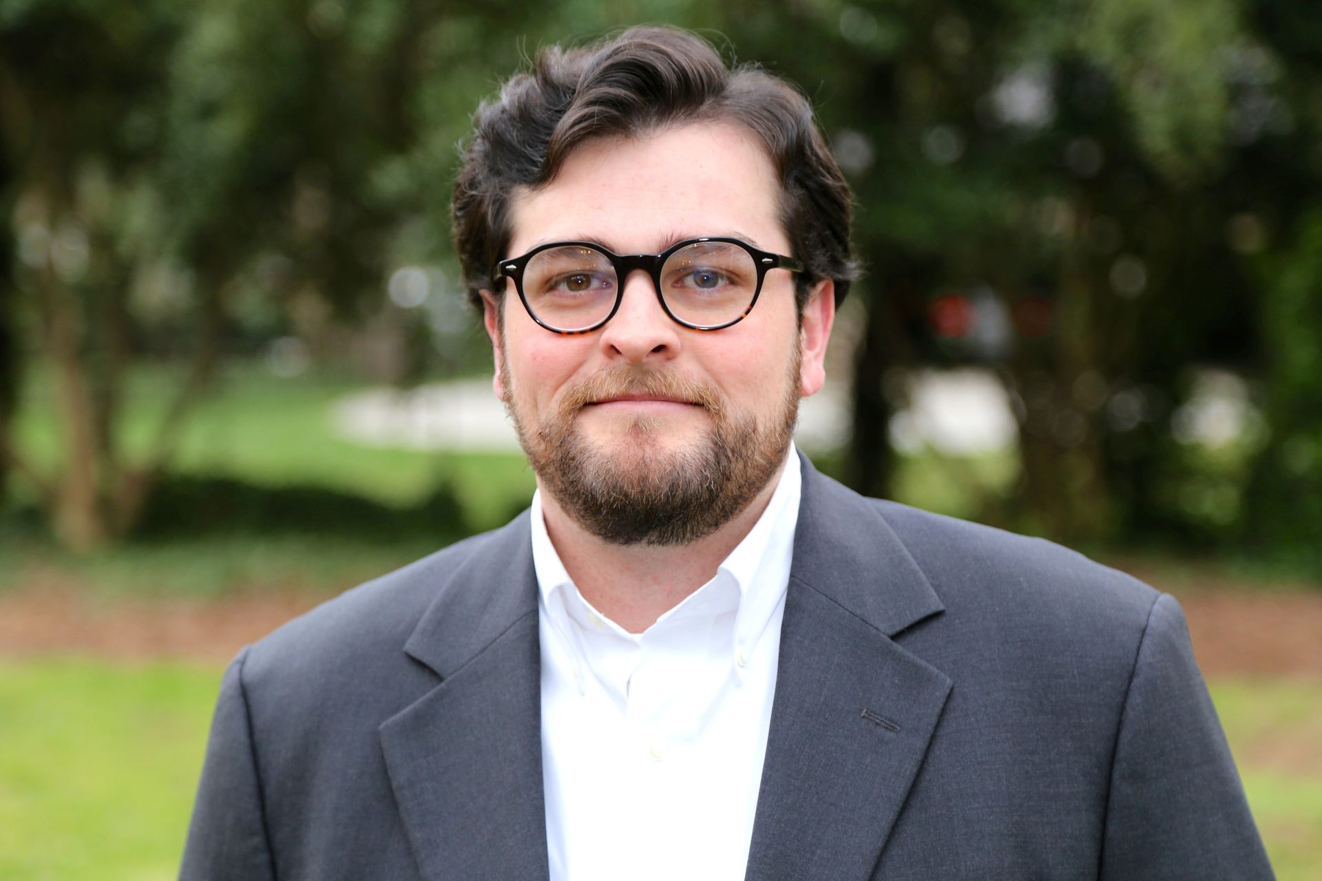 A man with a beard and glasses is wearing a suit and a white shirt.