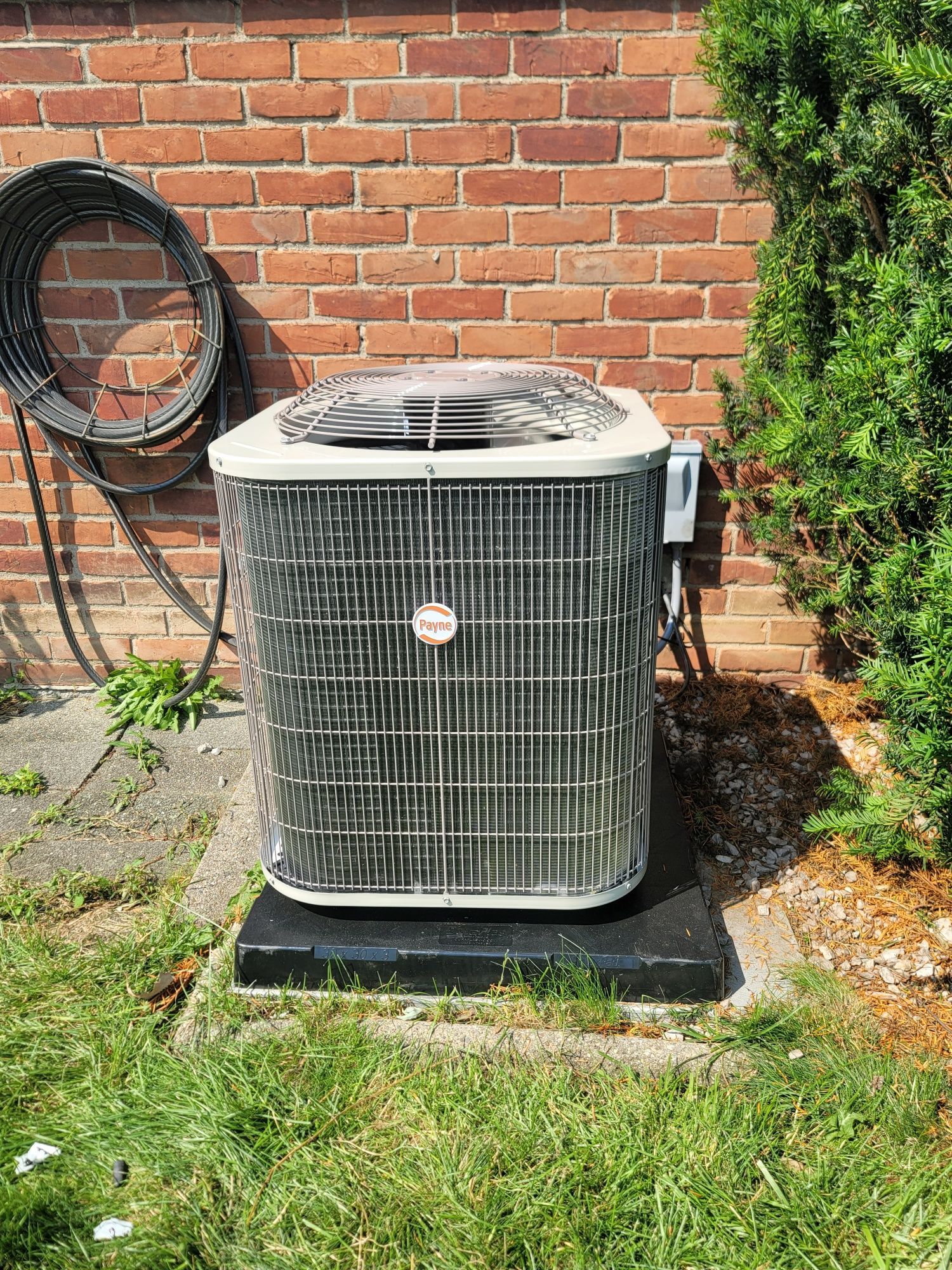 An air conditioner is sitting in the grass next to a brick wall.