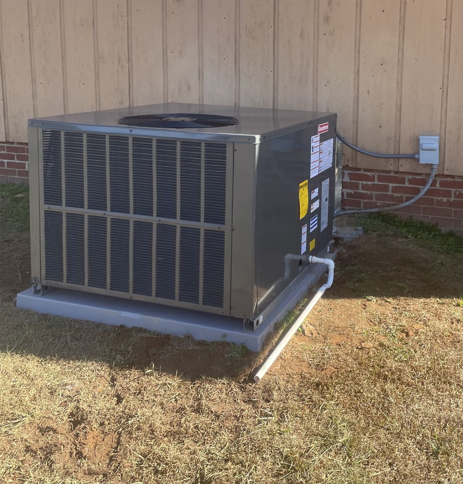 Outdoor air conditioning unit on a concrete pad beside a building. The unit is metallic gray with visible piping, set against a tan wooden wall.