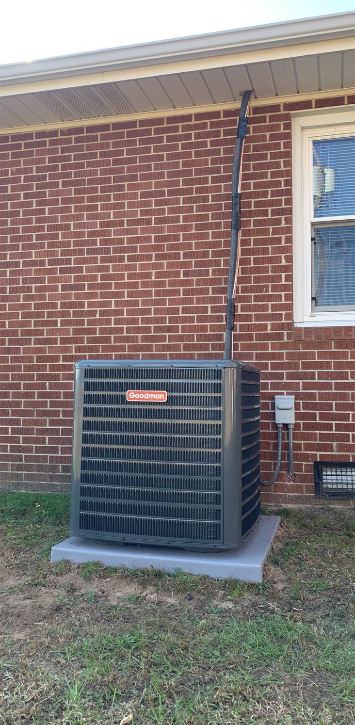 Outdoor air conditioning unit on a concrete pad next to a red brick house wall. A window and electrical cables are visible. Sunny day with grass surrounding the base.
