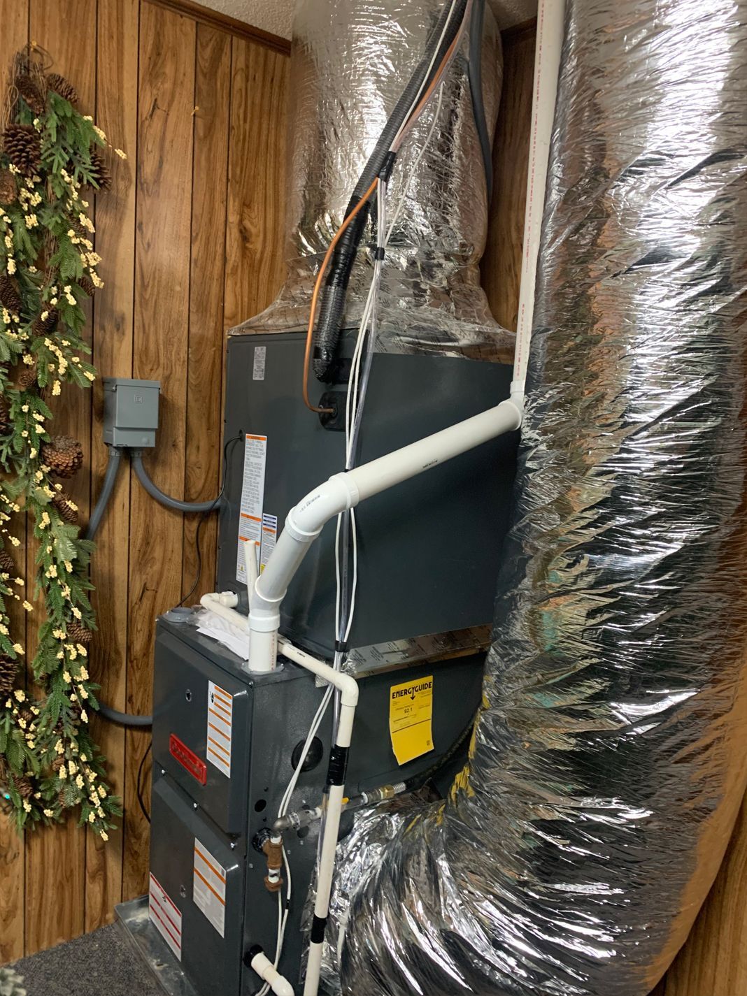 Image of a home furnace system with insulated ducts in a wood-paneled room. Pine cone and greenery decor adds a rustic touch. Functional and cozy setting.