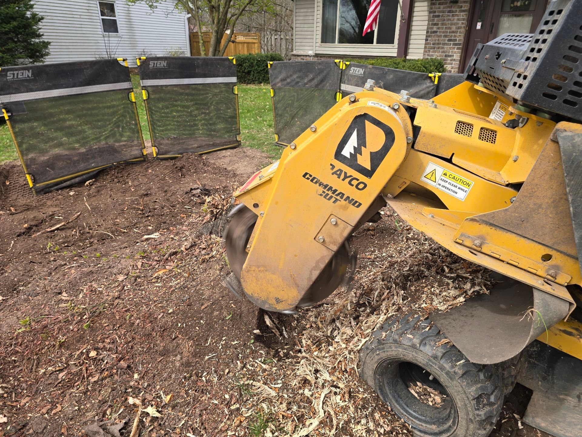 A yellow stump grinder is cutting a tree stump in front of a house.