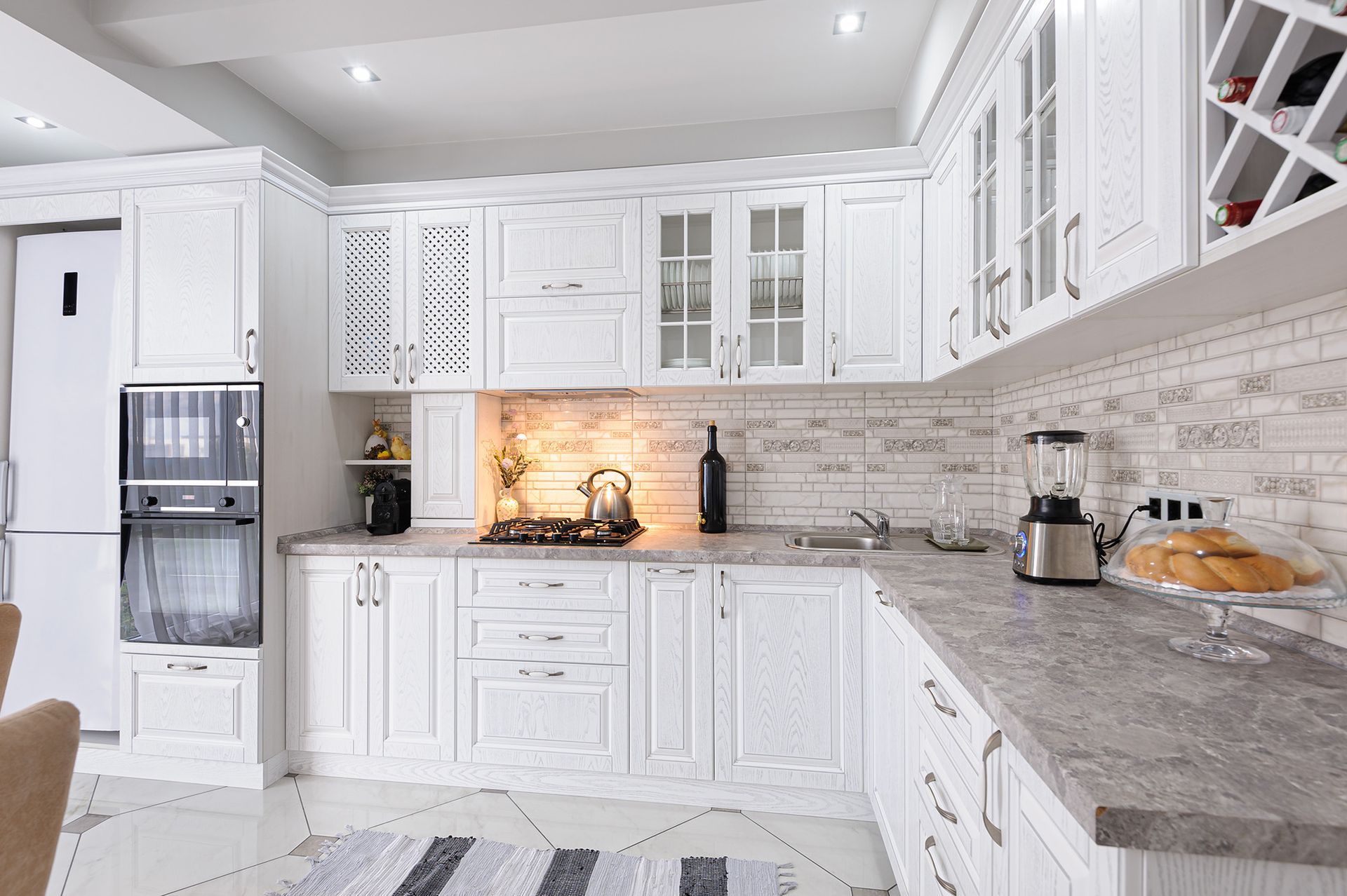 Bright white modern kitchen with marble counters, glass cabinets, and stainless steel appliances