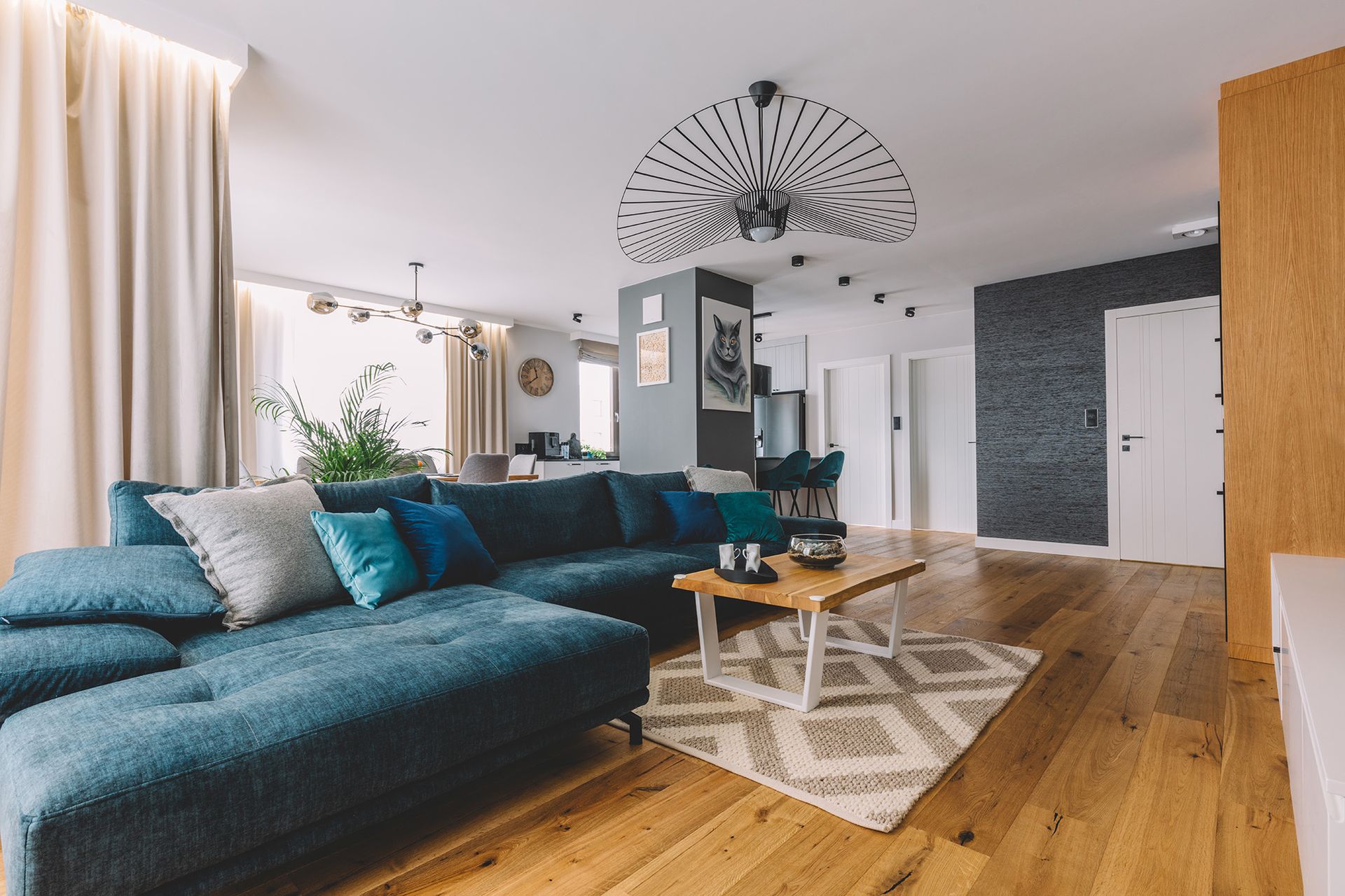 Modern living room with teal sectional sofa, wooden floors, patterned rug, and large windows