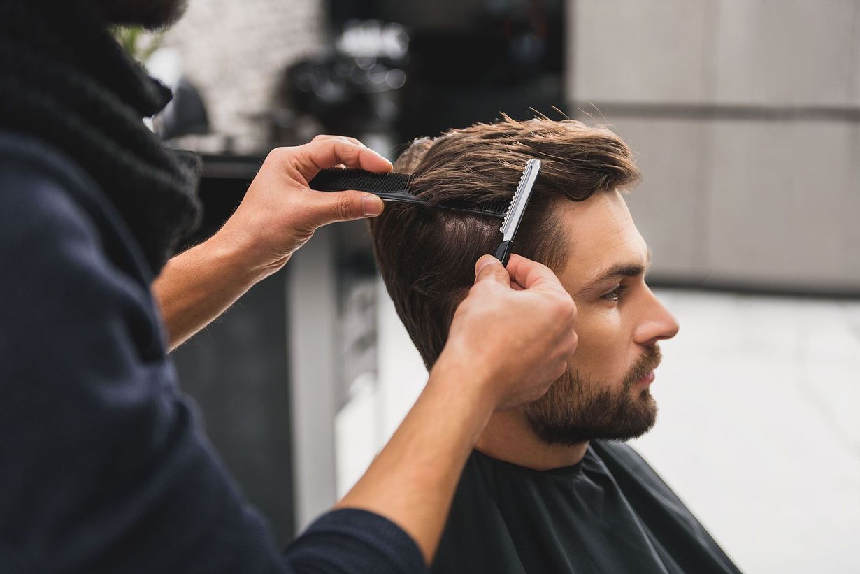 Barber cutting a man's hair with a comb and razor in a salon. The man has short, dark hair and a beard.