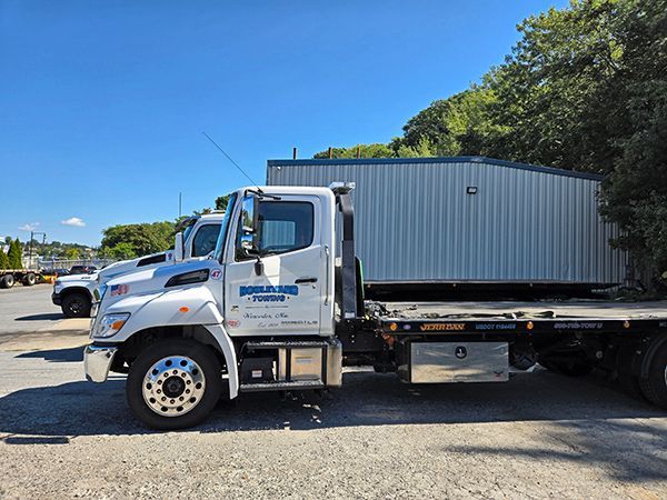 White tow truck hauling a gray metal storage unit on a sunny day.