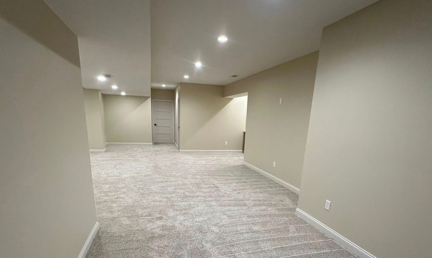 Empty beige basement room with gray carpet, recessed lights, and stairs in the back.