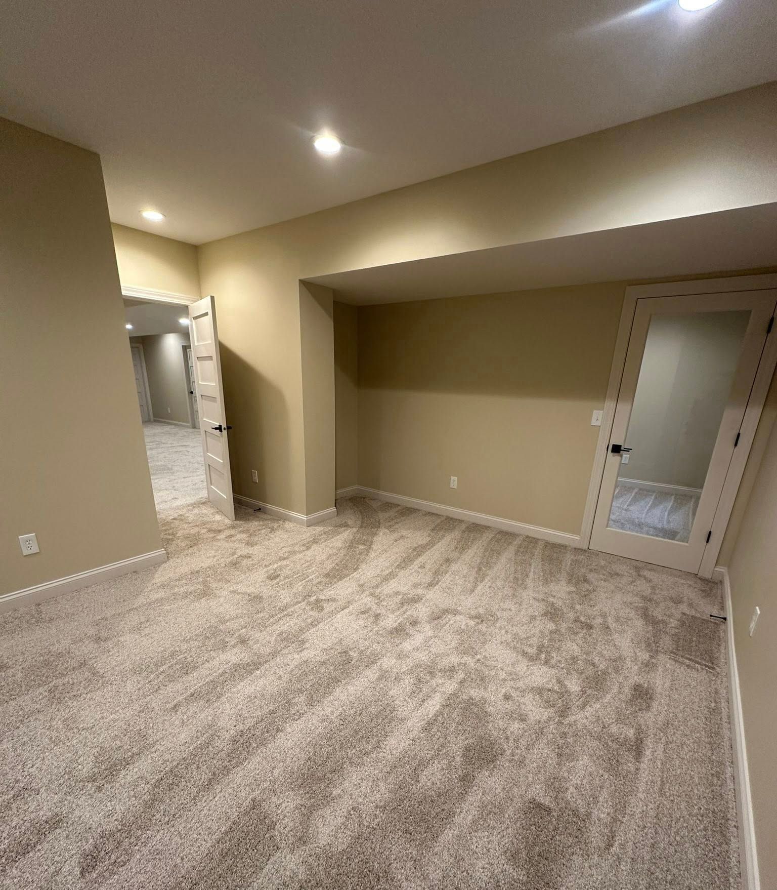 Empty beige carpeted room with recessed lights, alcove, and doorway to a hallway.