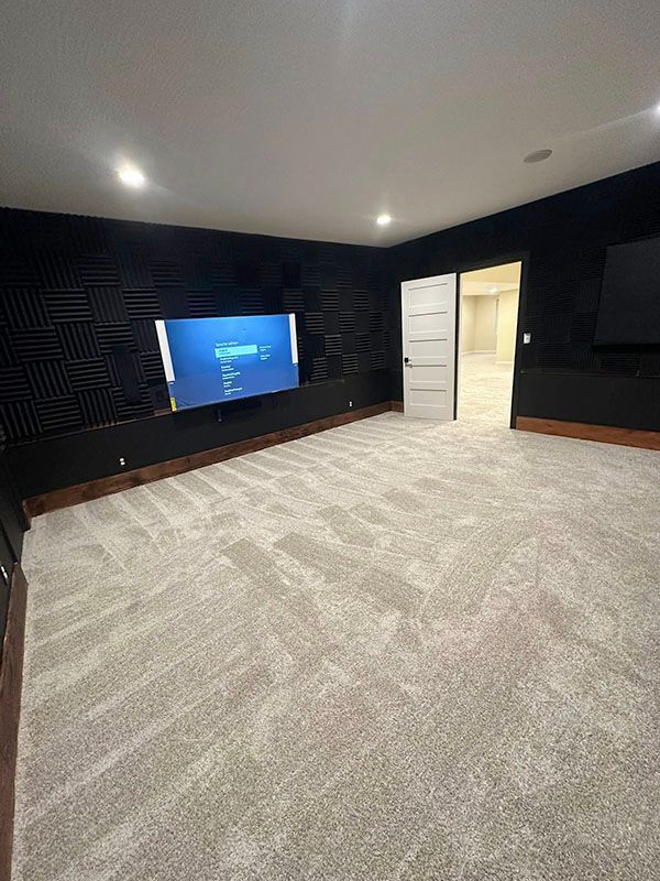 Empty carpeted media room with a wall-mounted TV, dark walls, and an open doorway to a lit hall