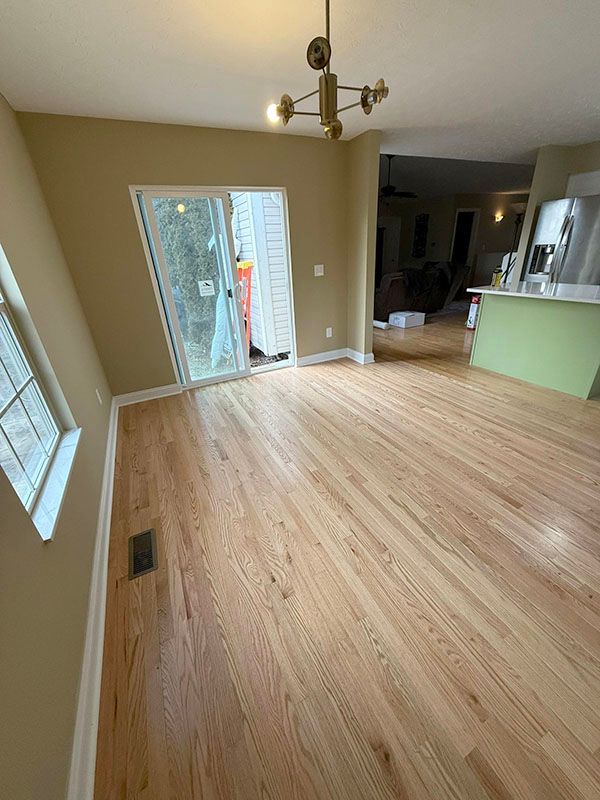 Empty living room with hardwood floors, beige walls, sliding glass door, and a view into the kitchen