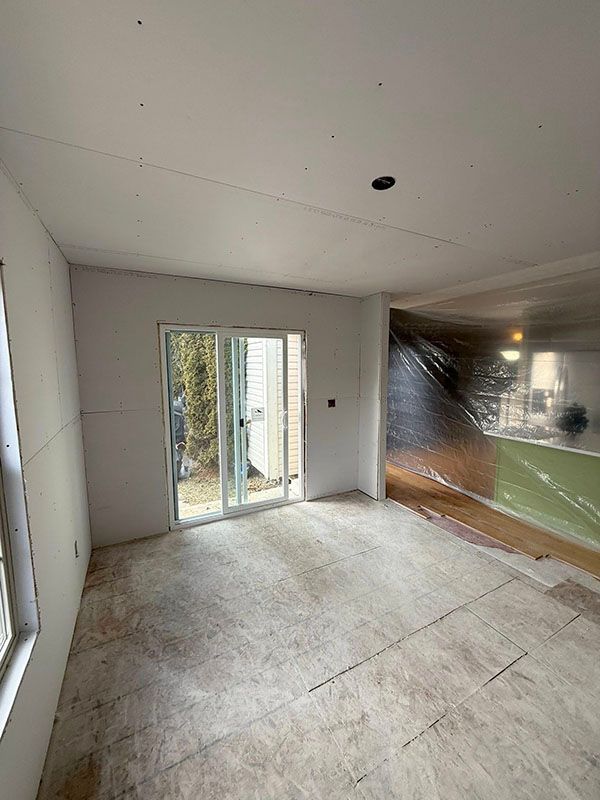 Empty unfinished room with white walls, concrete floor, sliding glass door, and plastic-covered opening to another space