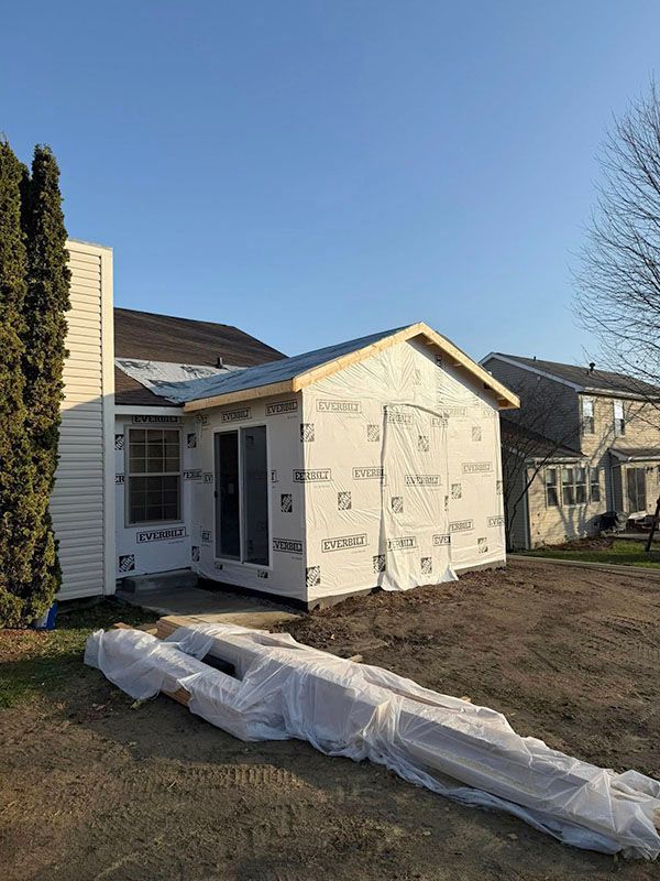 House exterior under renovation with white house wrap and plastic-covered materials on the lawn