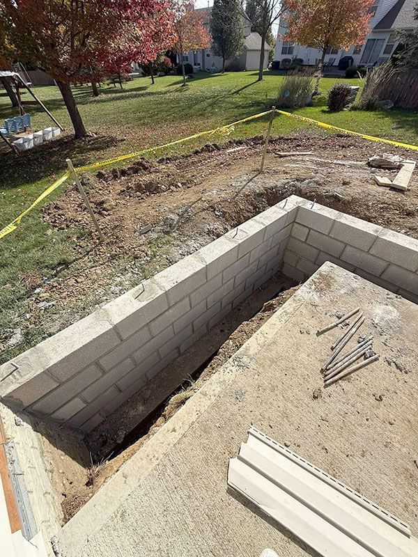 Partially built cinder block foundation with dirt floor and tools beside a grassy yard with autumn trees
