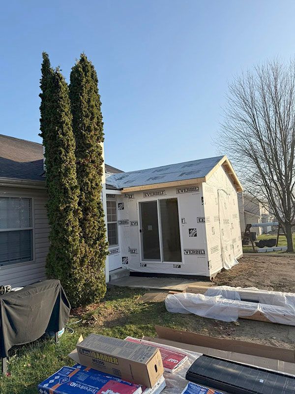 Small white house addition under construction beside a home, with materials and tall shrubs in the yard