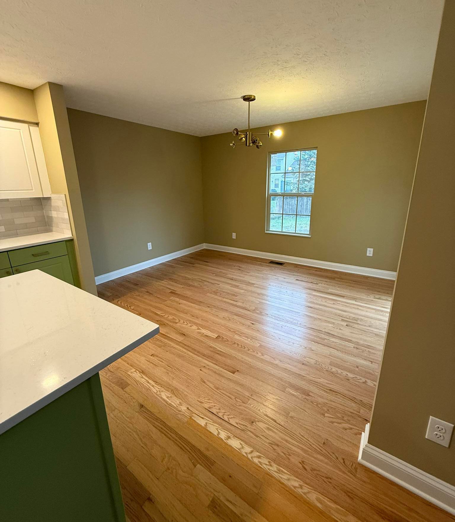 Empty kitchen-dining area with hardwood floors, beige walls, white trim, and a window.