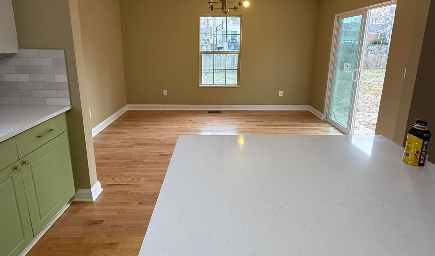 Empty kitchen with white island, green cabinets, wood floor, and a windowed back wall overlooking a patio doorway