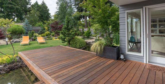 Wooden backyard deck beside a house, overlooking a landscaped garden with two orange chairs.