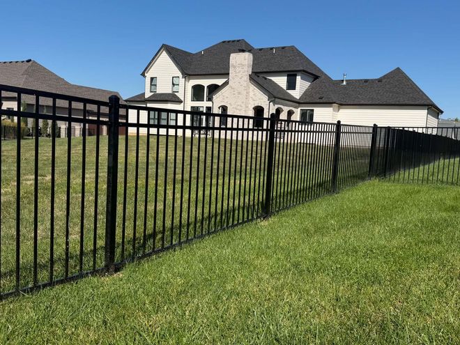 Black metal fence along a grassy yard in front of a large beige house under a clear blue sky