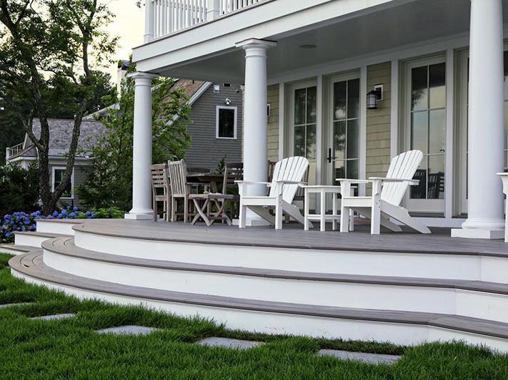 White porch with Adirondack chairs, round table, and steps on a gray house with landscaped lawn