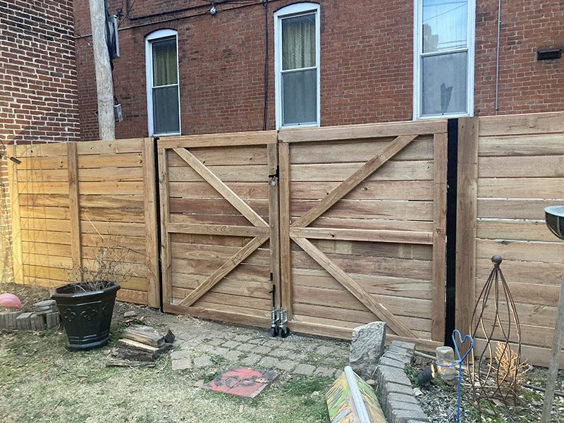 Wooden double gate with diagonal braces in a brick backyard, beside a fence and small patio area.
