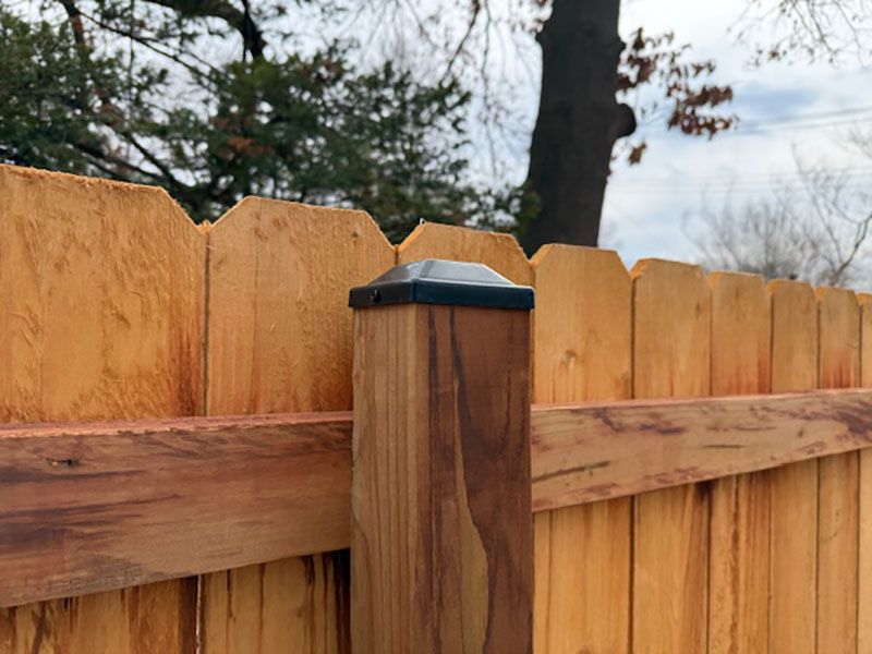 Wooden fence with a dark-topped post in a backyard, with trees and cloudy sky behind it