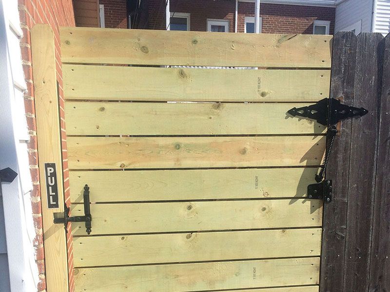 Light wood fence gate with black latch and a “PULL” sign, set between brick and dark wooden fences