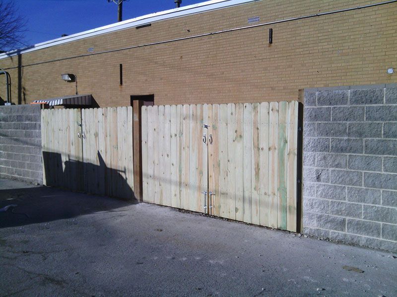 Weathered wooden gate in a brick wall between gray cinder block walls, beside an asphalt lot.