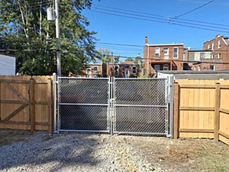 Chain-link gate between wooden fences, opening onto a gravel alley with brick buildings behind.