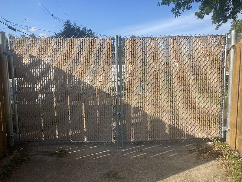 Closed chain-link gate in a fenced driveway, with sunlight and tree shadows overhead.