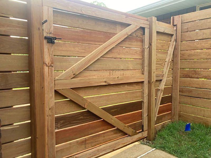 Wooden backyard gate with diagonal braces in a fenced yard, with grass and a small blue bucket nearby.