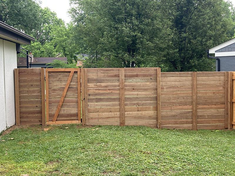 Wooden backyard fence with a gate, bordered by grass and trees.