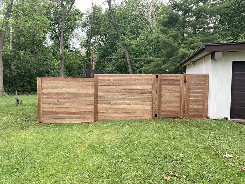 Wooden privacy fence beside a white building in a grassy yard with trees in the background