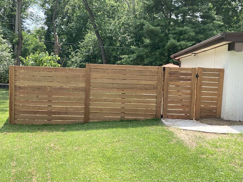 Wooden slat fence beside a house, with a gate and green lawn in front of trees.
