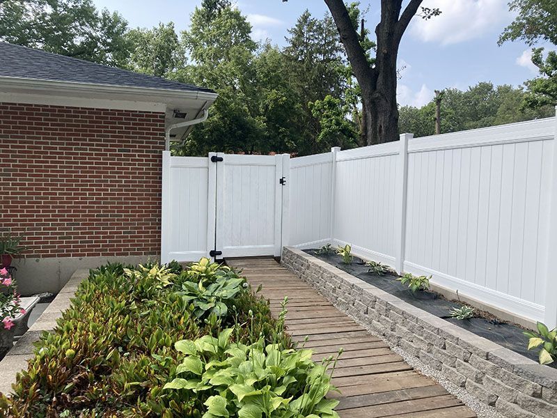 Backyard garden path beside white vinyl fence and brick house, with raised planter beds and trees.